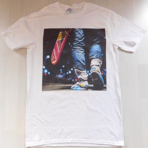 May include: White t-shirt with a printed image of a person wearing blue jeans and futuristic shoes, holding a pink hoverboard. The background is a blurry city scene at night.