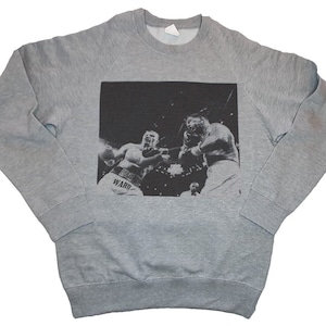 May include: A gray sweatshirt with a black and white image of two boxers in a boxing ring. The boxer on the left is wearing a white boxing glove and has the word "WARD" printed on his shorts. The boxer on the right is wearing a black boxing glove and has a black and white striped boxing robe.