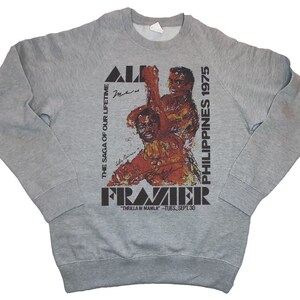 May include: Gray sweatshirt with a black and white graphic print of two boxers. The text "Ali Frazier" is printed in black letters with the text "The Saga of Our Lifetime" and "Philippines 1975" printed vertically on either side of the graphic. The text "Thrilla in Manila - Tues, Sept 30" is printed below the graphic.