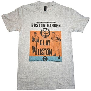 May include: A gray t-shirt with a vintage boxing poster print featuring Cassius Clay vs. Sonny Liston. The poster is for the World Heavyweight Championship at Boston Garden on May 25th, 1965. The text on the poster reads "World Heavyweight Championship, Boston Garden, May 25, Tues Eve, Champion Cassius Clay vs. Ex-Champion Sonny Liston, No TV."