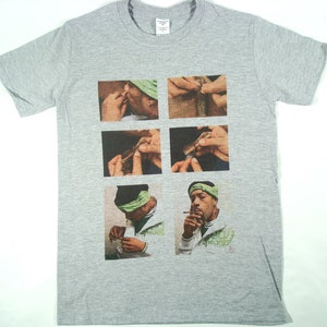 Puede incluir: Una camiseta gris con una imagen impresa de un hombre enrollando un cigarrillo. La imagen está dividida en cinco cuadrados, mostrando las manos del hombre enrollando el cigarrillo en diferentes etapas. El hombre lleva una bandana verde y una camisa blanca.