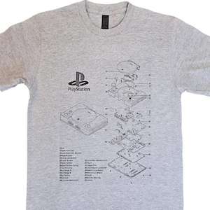 May include: A grey t-shirt with a detailed diagram of a PlayStation console and its internal components. The PlayStation logo is printed above the console diagram. The shirt has a crew neck and short sleeves.