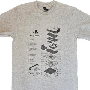 May include: A grey t-shirt displaying a detailed diagram of a PlayStation console's internal components. The PlayStation logo is printed above the diagram. The shirt includes a list of the parts.