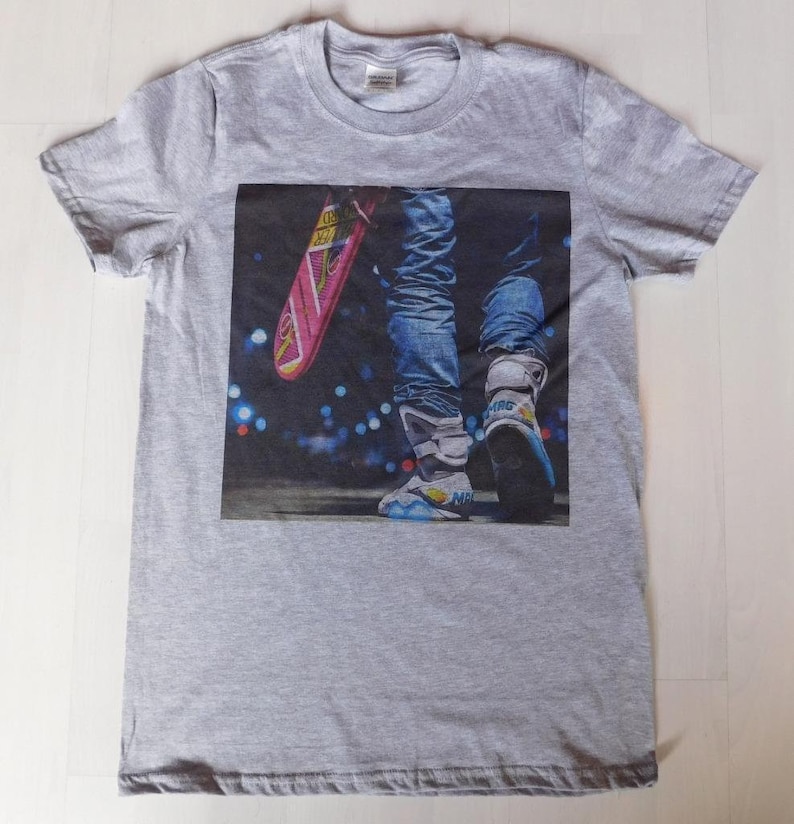 May include: A gray t-shirt with a printed image of a person wearing blue jeans and futuristic sneakers, holding a pink hoverboard.