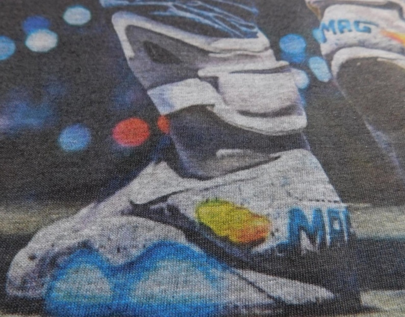 May include: A close-up of a gray and blue sneaker with the word "MAG" printed on the side. The sneaker is on a dark gray fabric background.