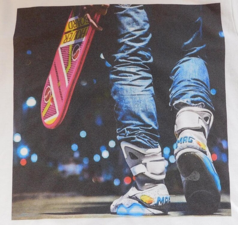 May include: A white t-shirt with a graphic print of a person wearing blue jeans and self-lacing sneakers. The person is holding a pink hoverboard with the text "Hoverboard" printed on it. The background is a blurry image of a city street at night with lights.