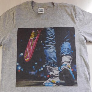 May include: Gray t-shirt with a graphic print of a person wearing blue jeans and futuristic sneakers holding a pink hoverboard. The background is a blurry city scene at night.