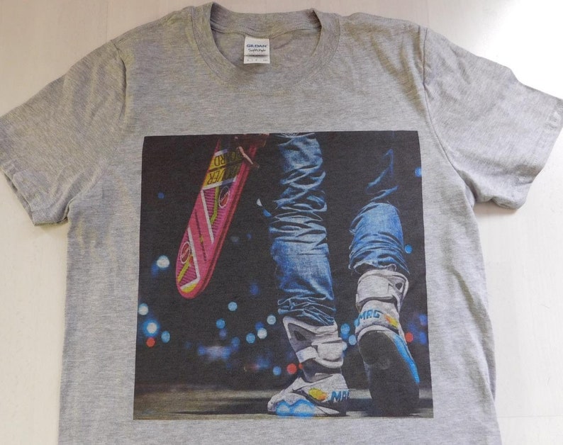 May include: Gray t-shirt with a graphic print of a person wearing blue jeans and futuristic sneakers holding a pink hoverboard. The background is a blurry city scene at night.