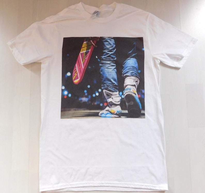 May include: White t-shirt with a printed image of a person wearing blue jeans and futuristic shoes, holding a pink hoverboard. The background is a blurry city scene at night.