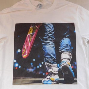 May include: White t-shirt with a printed image of a person wearing blue jeans and white sneakers holding a pink and blue hoverboard. The background is a blurry image of city lights.