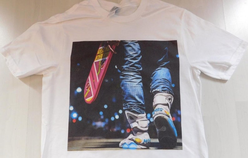 May include: White t-shirt with a printed image of a person wearing blue jeans and white sneakers holding a pink and blue hoverboard. The background is a blurry image of city lights.