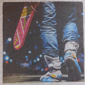 May include: A grey t-shirt with a graphic print of a person wearing blue jeans and white sneakers with a pink hoverboard. The person is standing on a dark background with blurry lights.