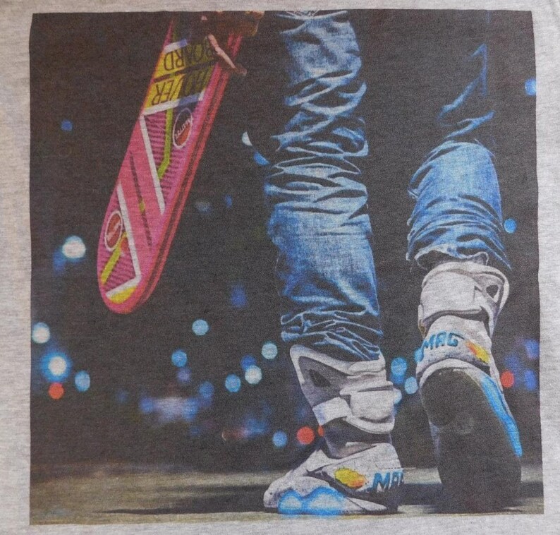 May include: A grey t-shirt with a graphic print of a person wearing blue jeans and white sneakers with a pink hoverboard. The person is standing on a dark background with blurry lights.