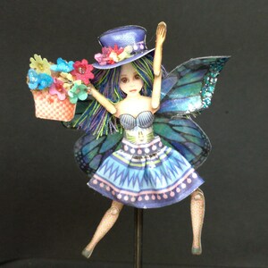 Könnte beinhalten: Eine blaue und grüne Feenpuppe mit einem blauen Hut, einem Blumenkorb und Schmetterlingsflügeln. Die Puppe trägt ein blaues und weißes Kleid mit rosa Akzenten.