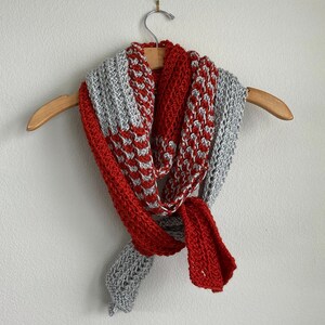 May include: A gray and red crocheted scarf hanging on a wooden hanger. The scarf is made with a simple stitch pattern and has a long, flowing shape.