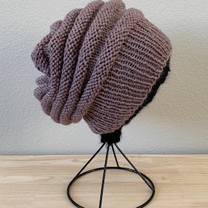May include: A brown and black knitted beanie hat with a slouchy design. The hat is displayed on a black metal stand.