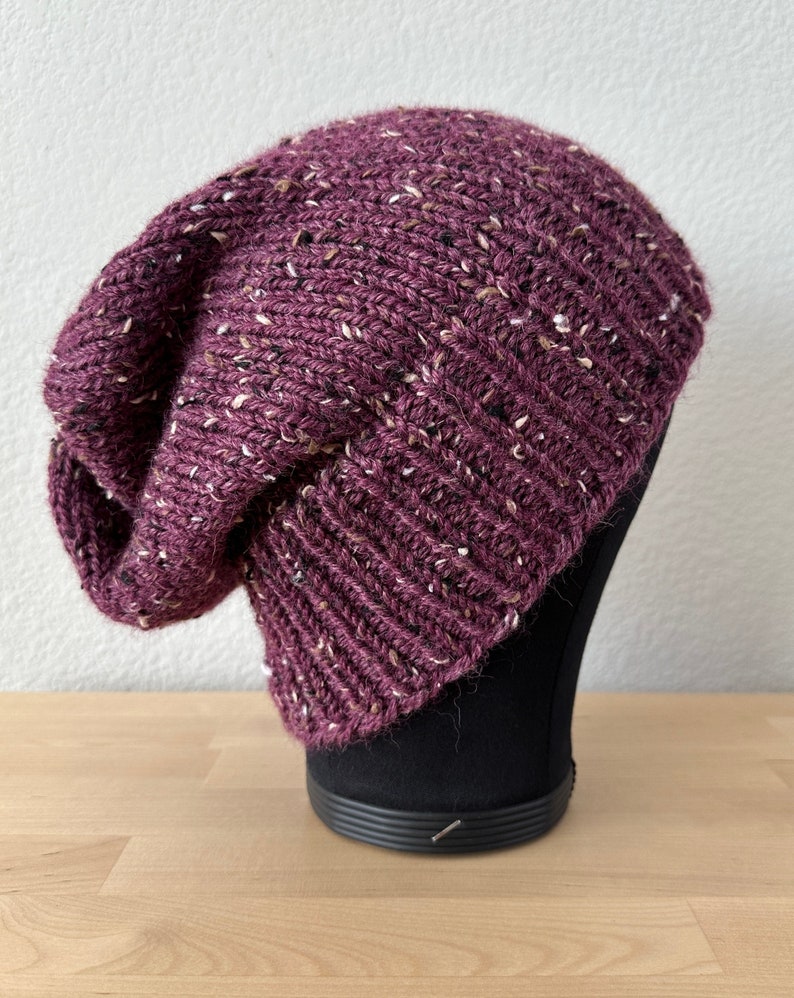 May include: A slouchy beanie hat in a deep burgundy color with flecks of white and brown throughout the knit.