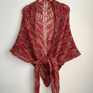 May include: A triangular shawl featuring an openwork knit pattern in a variegated blend of deep red and metallic gold yarn. The garment is draped to show a central lace design and long, pointed ends that are tied together at the front center.