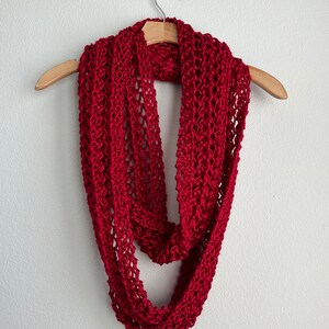 May include: A red crocheted infinity scarf hanging on a wooden hanger against a white wall.