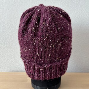 May include: A purple knitted beanie with white flecks throughout. The beanie is sitting on a black display stand.