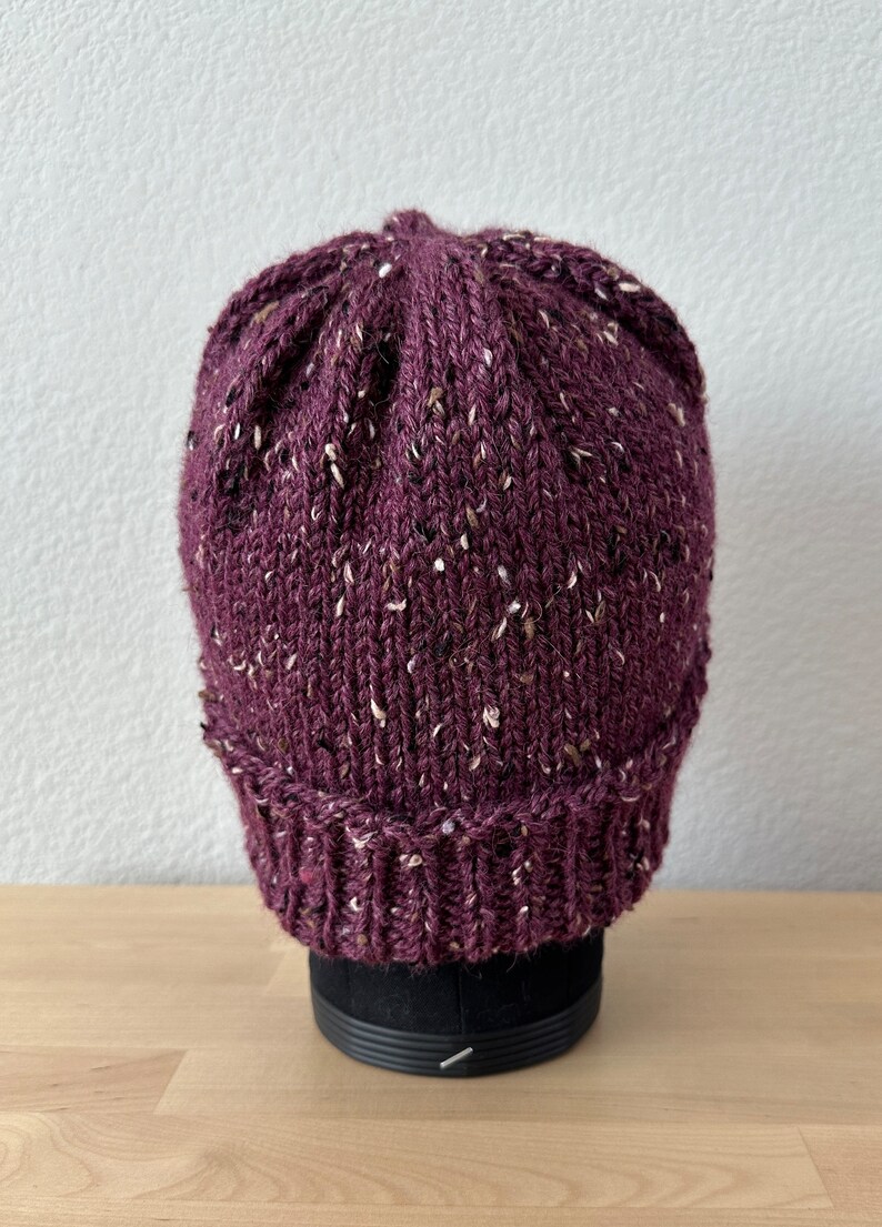 May include: A purple knitted beanie with white flecks throughout. The beanie is sitting on a black display stand.