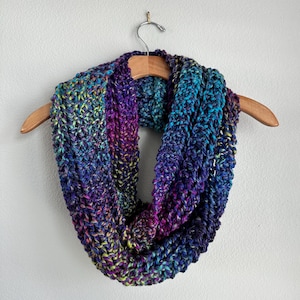 May include: A chunky knit infinity scarf featuring a variegated yarn pattern with vibrant shades of deep purple, royal blue, teal, and hints of lime green. The scarf is constructed with a thick, textured stitch pattern and hangs in a circular loop.