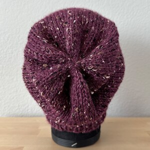 May include: A purple knitted beanie with a speckled pattern. The beanie is on a black display stand.