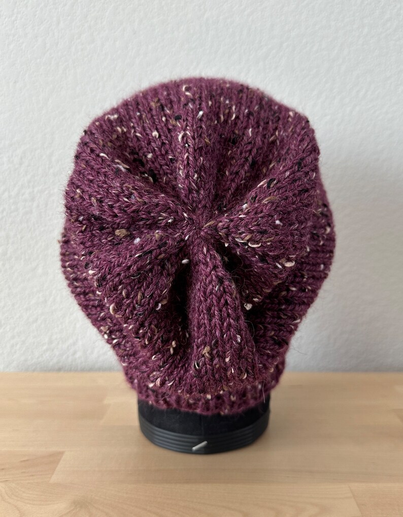 May include: A purple knitted beanie with a speckled pattern. The beanie is on a black display stand.