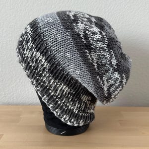 May include: A gray, white, and black knit beanie hat. The hat features a geometric pattern and is displayed on a black form. The beanie is made of a soft, textured material, perfect for cold weather. The hat is a stylish accessory.