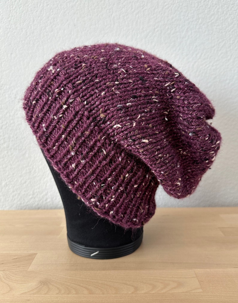 May include: A purple knitted beanie with white flecks throughout. The beanie is slouchy and has a ribbed texture.