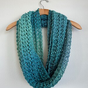 Peut inclure: Une écharpe infinie en crochet dans un dégradé de couleurs turquoise et vertes. L'écharpe est suspendue à un cintre en bois.