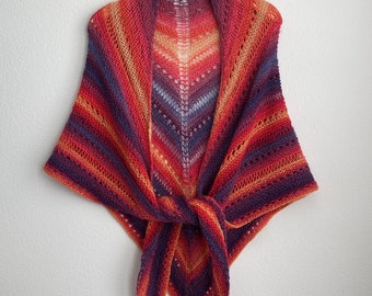 Puede incluir: Un chal triangular de punto con un patrón de encaje en tonos naranja, rojo, morado y azul. El chal está anudado en el centro.