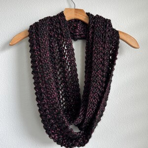 May include: A hand-knitted infinity scarf in a dark color palette of black and burgundy. The chunky knit scarf is displayed on a wooden hanger against a white background. The scarf's texture and color variations are clearly visible.