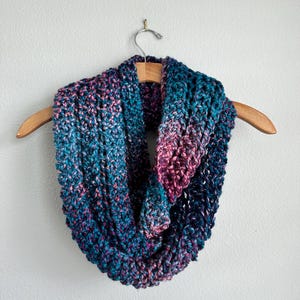 May include: A chunky knit infinity scarf featuring a variegated color palette of deep navy blue, vibrant teal, and magenta pink. The yarn has a thick, textured appearance with a consistent loop stitch pattern, draped over a wooden clothes hanger.