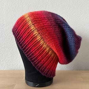Puede incluir: Un gorro tejido a mano con un degradado de colores, que va del rojo al naranja y luego al azul. El gorro se muestra sobre una cabeza de maniquí negra, mostrando su textura acanalada y su estilo holgado. El gorro está hecho de hilo suave.