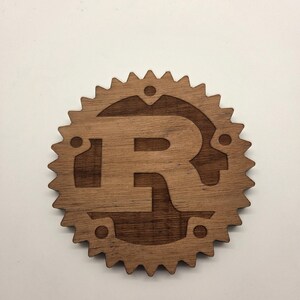 Könnte beinhalten: Ein Holz-Emblem mit dem Buchstaben "R" in einem Zahnrad-Design. Das Emblem besteht aus hellbraunem Holz mit dunklerem braunem Innenraum und hat eine kreisförmige Form mit spitzen Kanten. Das Design ist wahrscheinlich ein Logo oder Symbol.