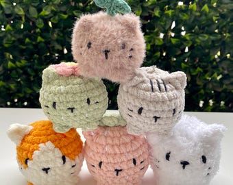 May include: A stack of six crocheted cat-shaped toys in various colors: orange and white, light green, pink, gray, white, and a fluffy brown with a green leaf. Each cat has embroidered facial features.