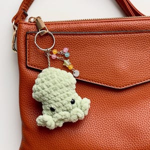 May include: A light green crocheted octopus keychain with black eyes and a silver keyring. The keychain has a beaded charm with pink, yellow, and blue beads, and a star charm. The keychain is attached to a brown leather purse.