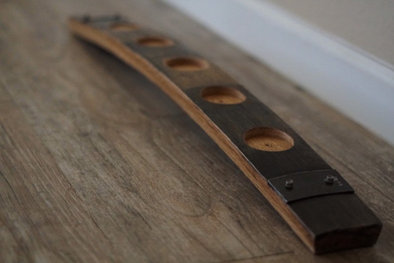 Op de afbeelding: Een houten kandelaar gemaakt van een gebogen stuk van een vat. Het heeft vijf ronde uitsparingen voor kaarsen en donkere metalen beslag. Het hout heeft een natuurlijke, warme tint.
