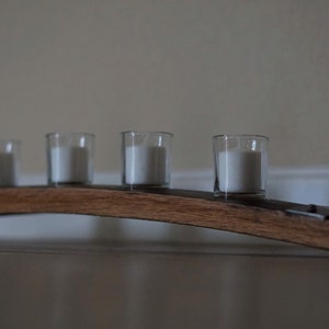 Op de afbeelding: Een houten kandelaar met vijf heldere glazen votiefhouders, elk met een witte kaars. De houder is gemaakt van gebogen, donker gebeitst hout en is vastgezet met metalen beugels. De kaarsen zijn niet aangestoken.