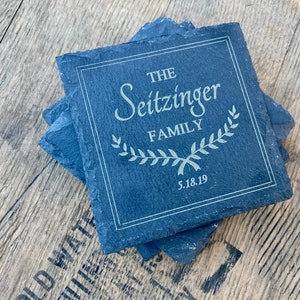 Op de afbeelding: Set van vier zwarte leisteen onderzetters met een witte rand en de tekst "THE Seitzinger FAMILY 5.18.19" in witte letters met een groen kransontwerp.