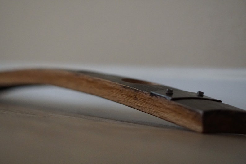 Op de afbeelding: Een gebogen houten staaf van een vat, met natuurlijke houtnerf en donkere metalen beslag. De staaf heeft een gat en maakt waarschijnlijk deel uit van een wijn- of whiskyvat.