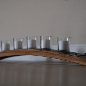 Op de afbeelding: Een houten kandelaar met vijf doorzichtige glazen votiefkaarsen, elk met een witte kaars. De houder is gemaakt van een gebogen stuk hout, waarschijnlijk van een vat, en heeft metalen accenten. De kaarsen zijn niet aangestoken.