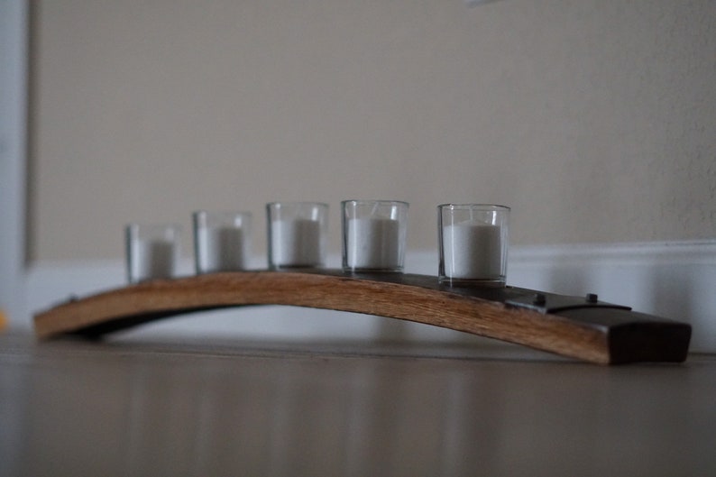 Op de afbeelding: Een houten kandelaar met vijf doorzichtige glazen votiefkaarsen, elk met een witte kaars. De houder is gemaakt van een gebogen stuk hout, waarschijnlijk van een vat, en heeft metalen accenten. De kaarsen zijn niet aangestoken.