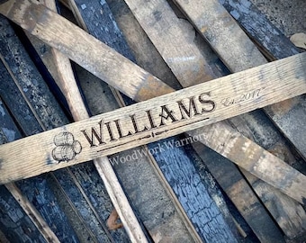 May include: A wooden sign with the name "WILLIAMS" engraved in a decorative font, along with "Est. 2017". The sign also features an illustration of three barrels. The sign is made of weathered wood.