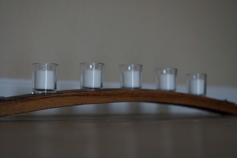 Op de afbeelding: Een houten kandelaar met vijf doorzichtige glazen votiefhouders, elk met een witte kaars. De houder heeft een gebogen vorm en is gemaakt van donkerbruin hout. De kaarsen zijn gelijkmatig verdeeld.