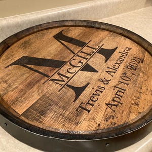 Op de afbeelding: Een ronde houten tondeksel met een donkere metalen rand. Het deksel is gegraveerd met de naam "McGill" en de namen "Trevis & Alexandria" samen met de datum "April 10th, 2021". Het hout heeft een natuurlijke, verouderde uitstraling.