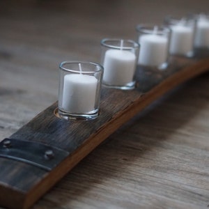 Op de afbeelding: Een rustieke kandelaar gemaakt van een gebogen stuk hout, met vijf heldere glazen votiefhouders die elk een witte kaars bevatten. Het hout heeft een donkere, verweerde uitstraling, met metalen accenten. Een decoratief item voor in huis.