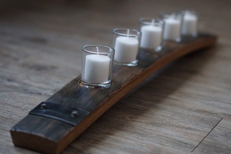 Op de afbeelding: Een rustieke kandelaar gemaakt van een gebogen stuk hout, met vijf heldere glazen votiefhouders die elk een witte kaars bevatten. Het hout heeft een donkere, verweerde uitstraling, met metalen accenten. Een decoratief item voor in huis.