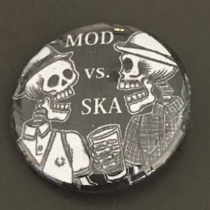 May include: Black and white button with two skeletons wearing hats. The skeleton on the left is holding a glass of beer. The text on the button reads "MOD VS. SKA".